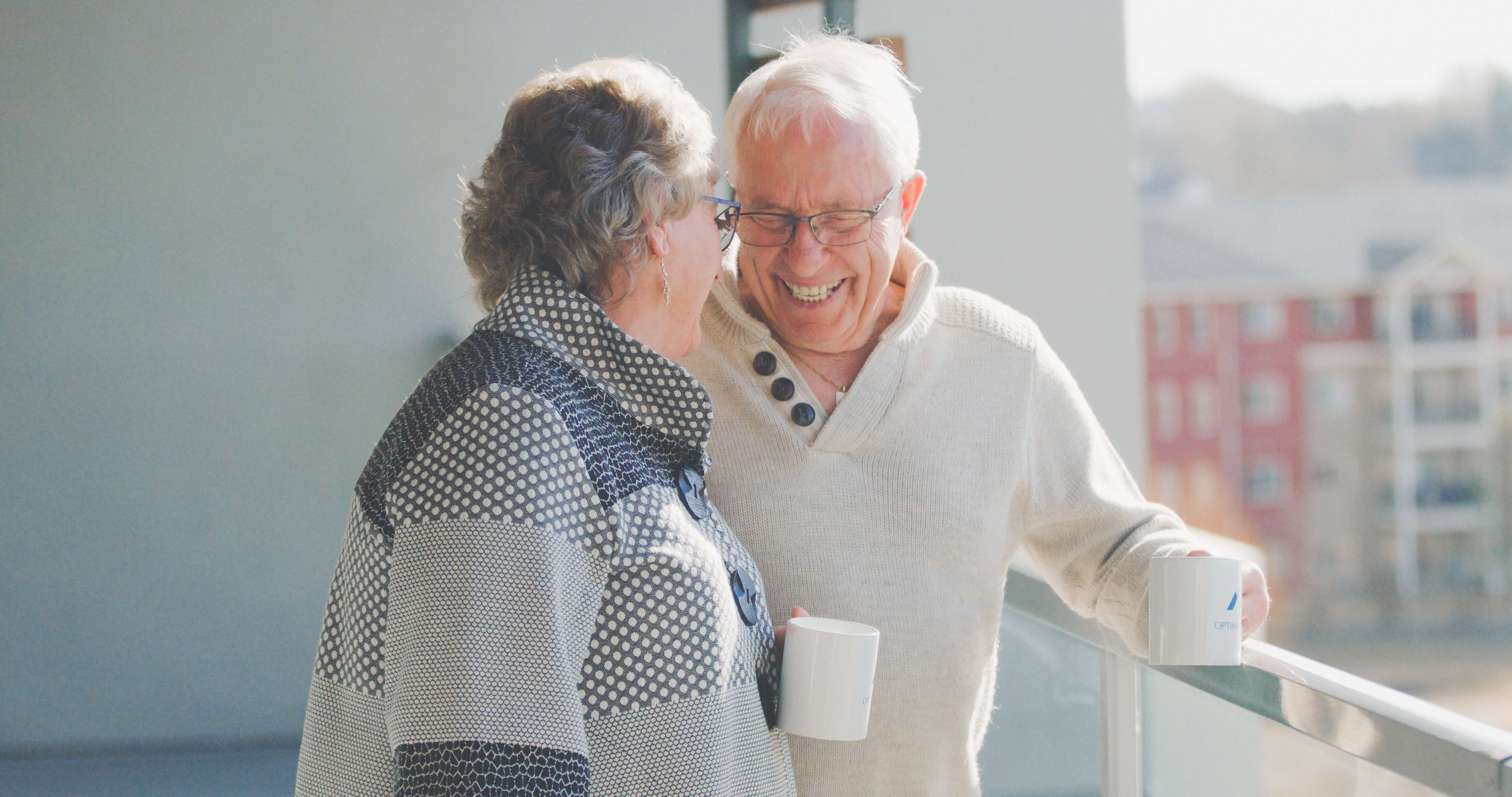 An senior couple laughing, holding coffee mugs on a balcony