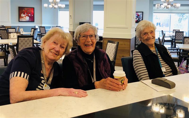 Three senior women seated together smiling. One of the ladies has a cup of coffee.