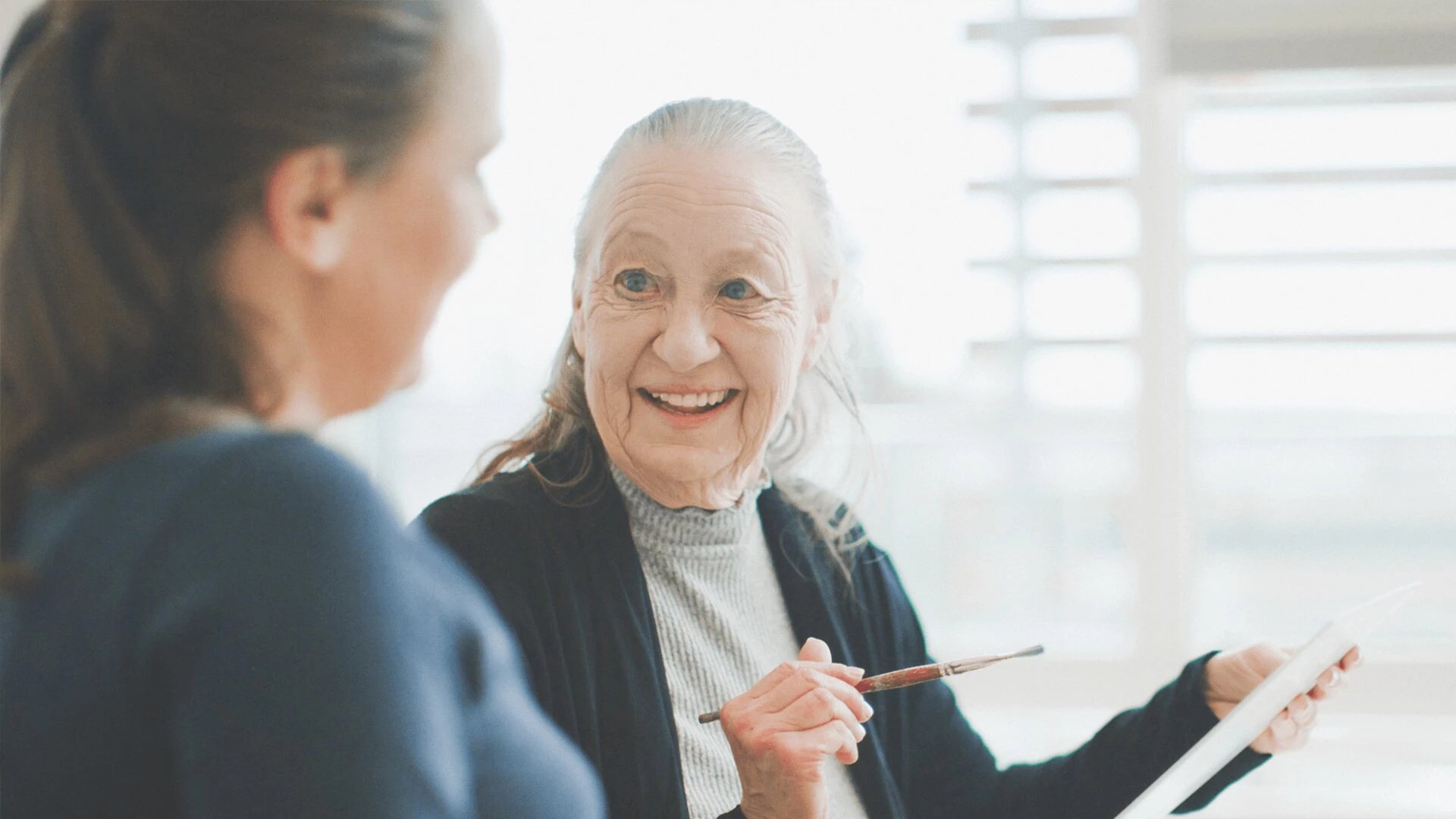 A senior woman holding a paintbrush and canvas and talking to a young woman